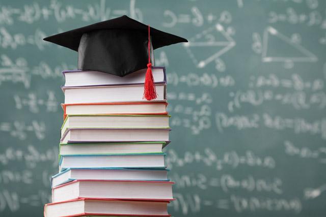 a stack of books in front of a chalk board with a graduation cap on top